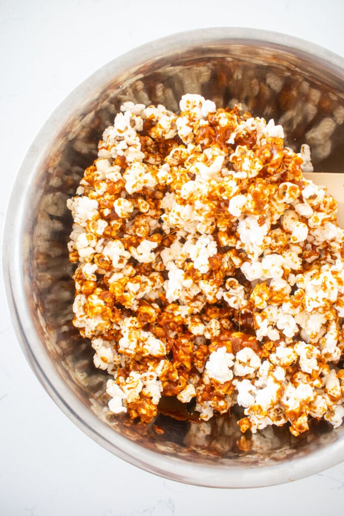 caramel being stirred into popcorn in a big mixing bowl on a marble counter.