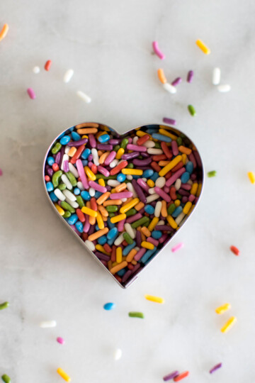 rainbow sprinkles inside a heart cookie cutter on a white marble background.