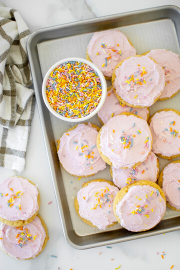 soft sugar cookies with pink frosting and rainbow sprinkles on a baking sheet on a white marble counter.