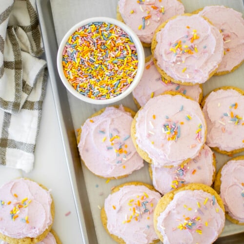 soft sugar cookies with pink frosting and rainbow sprinkles on a baking sheet on a white marble counter.