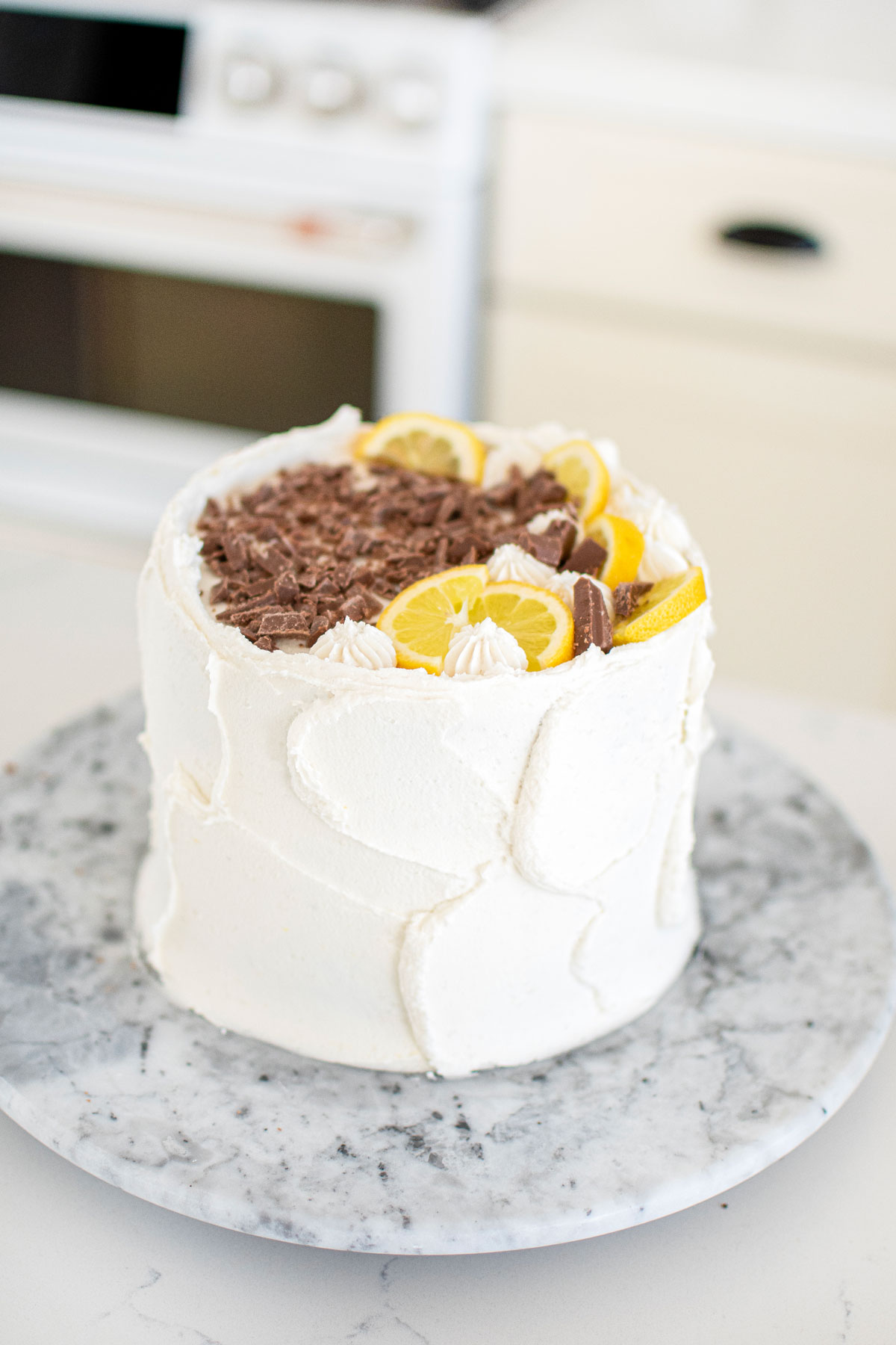 chocolate lemon cake with chocolate shavings and lemon slices on top sitting on a marble cake spinner on kitchen counter.