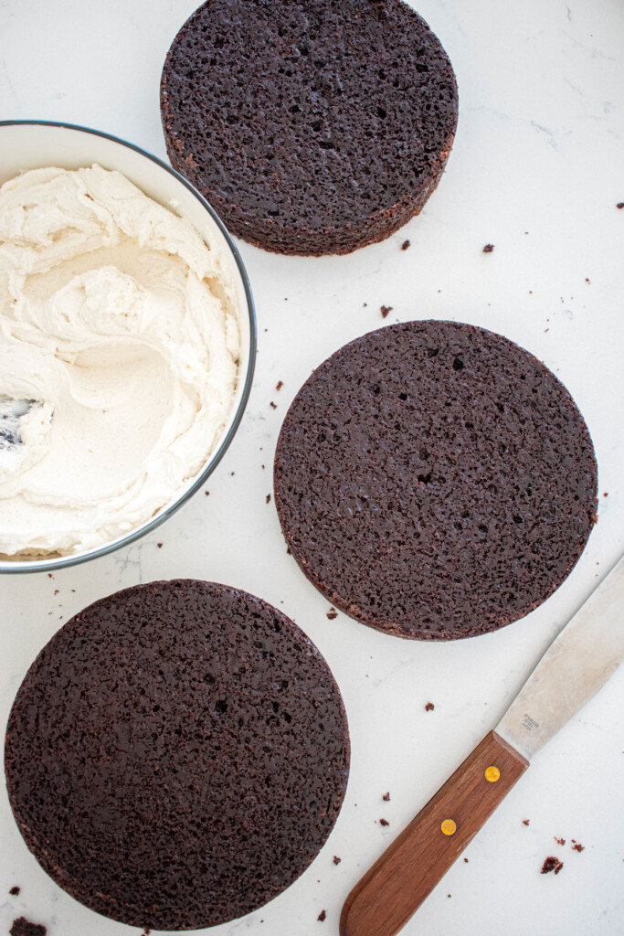 chocolate cake layers and vanilla frosting in a bowl with a cake spatula on a white marble counter.