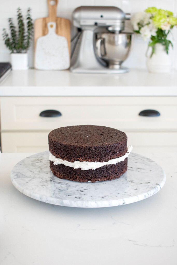 chocolate cake with vanilla frosting being stacked on a cake spinner on a white marble counter.