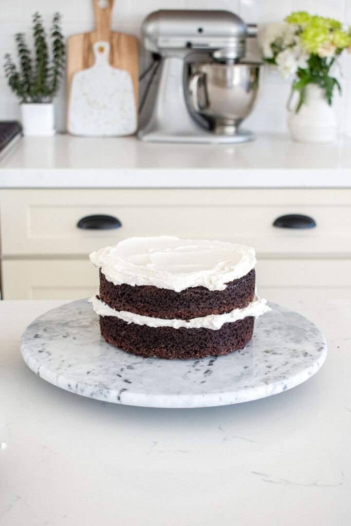 chocolate cake with vanilla frosting being stacked on a cake spinner on a white marble counter.