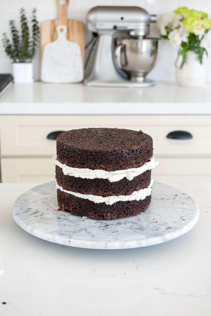 chocolate cake with vanilla frosting being stacked on a cake spinner on a white marble counter.