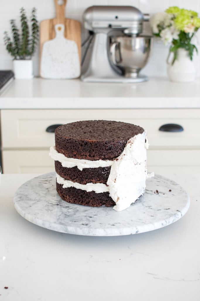 chocolate cake with vanilla frosting being crumb coated on a cake spinner on a white marble counter.