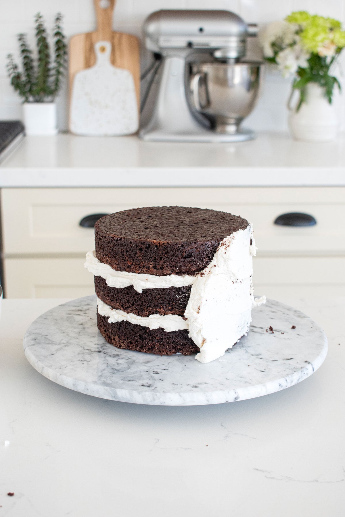 chocolate cake with vanilla frosting being crumb coated on a cake spinner on a white marble counter.