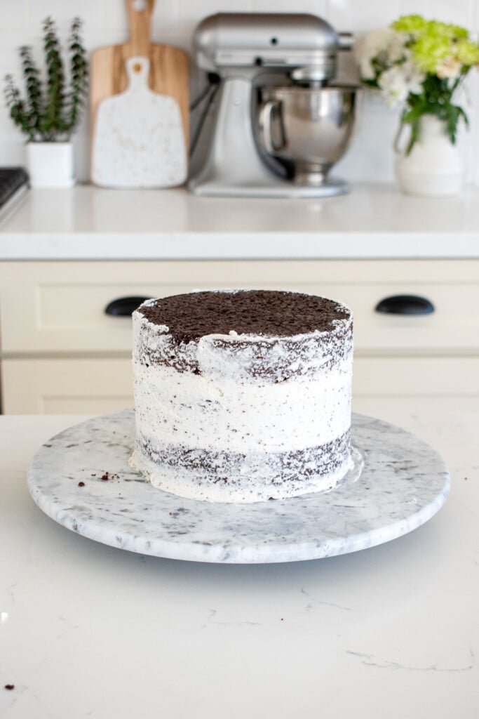 chocolate cake with vanilla frosting being crumb coated on a cake spinner on a white marble counter.