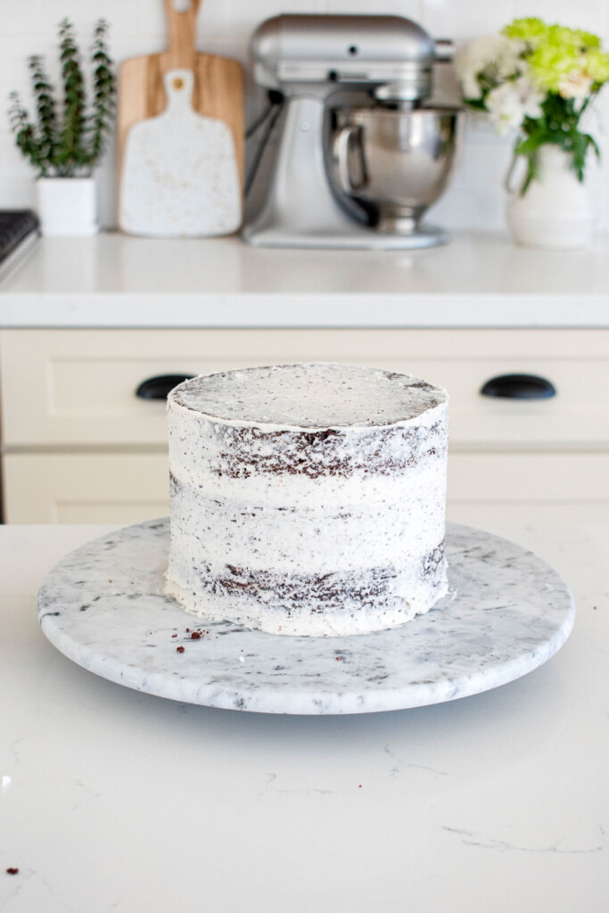 chocolate cake with vanilla frosting being crumb coated on a cake spinner on a white marble counter.