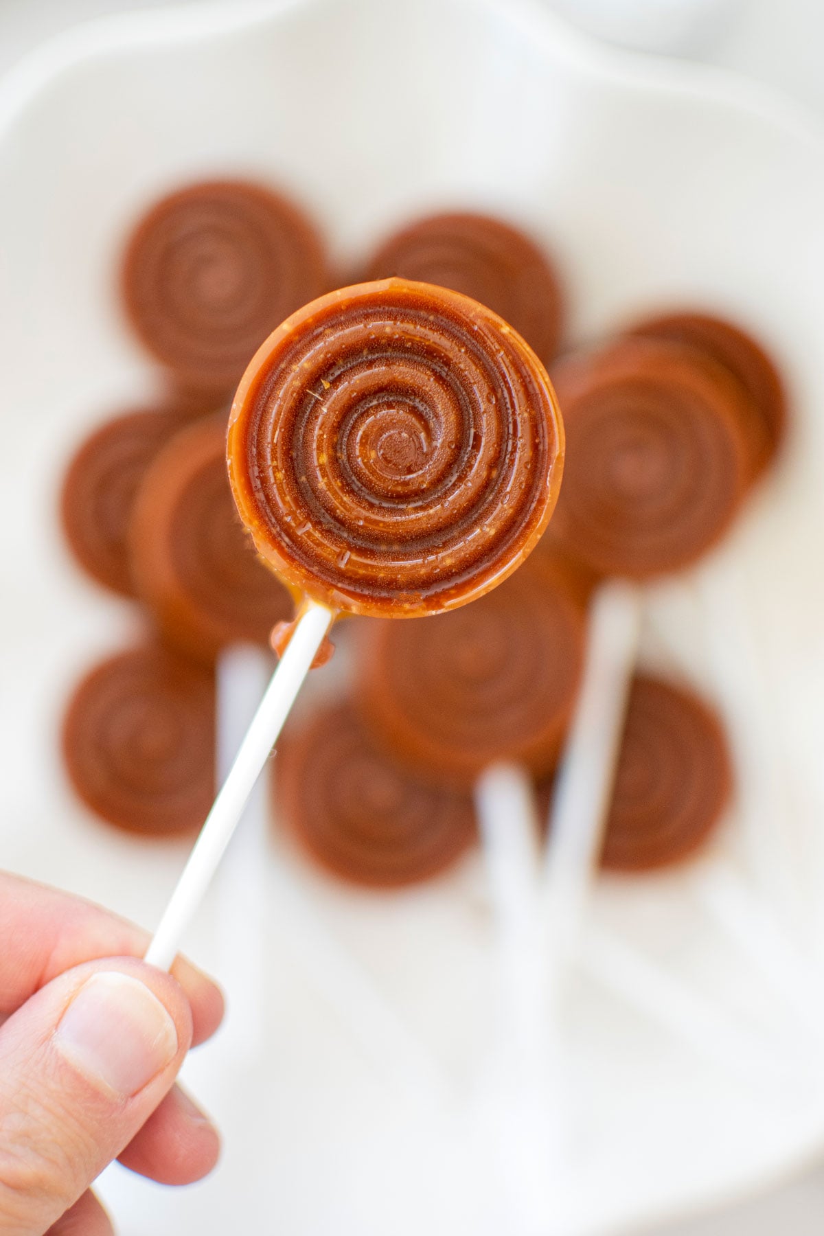 woman holding a caramel lollipop up over a plate of more caramel lollipops.