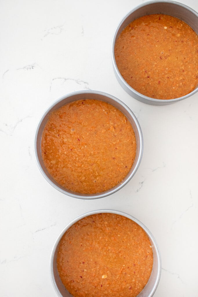 honey apple carrot cake batter in 3 cake pans on a white marble counter.