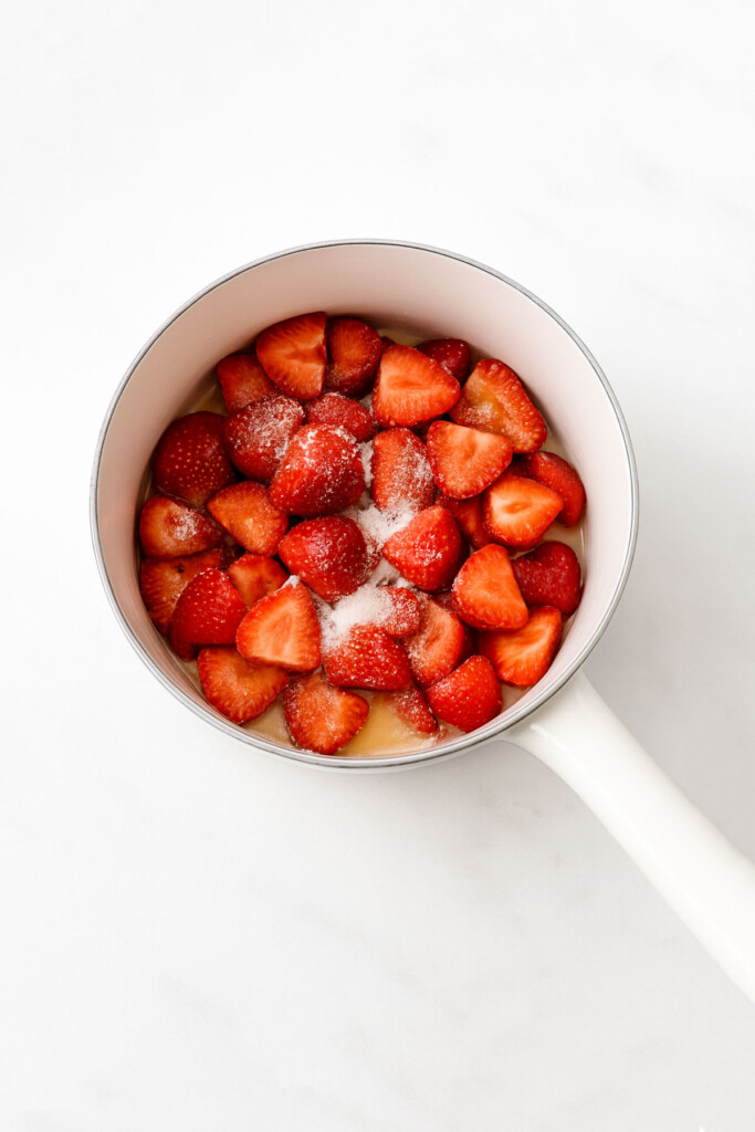 sliced strawberries, cane sugar and lemon juice in a white pot on a marble counter.