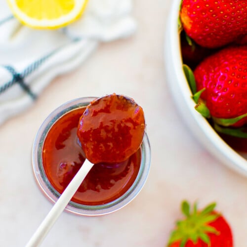 organic strawberry jam in a glass jar with a spoon on a table with fresh strawberries and a lemon.
