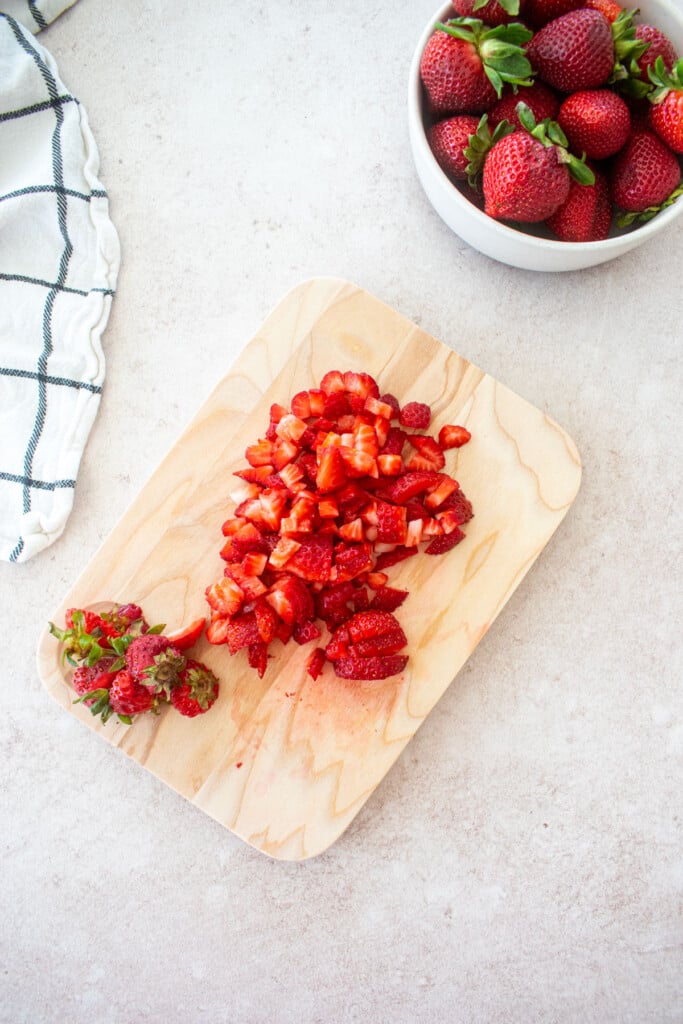 wood cutting board with chopped strawberries and a bowl of fresh strawberries on the counter next to it.