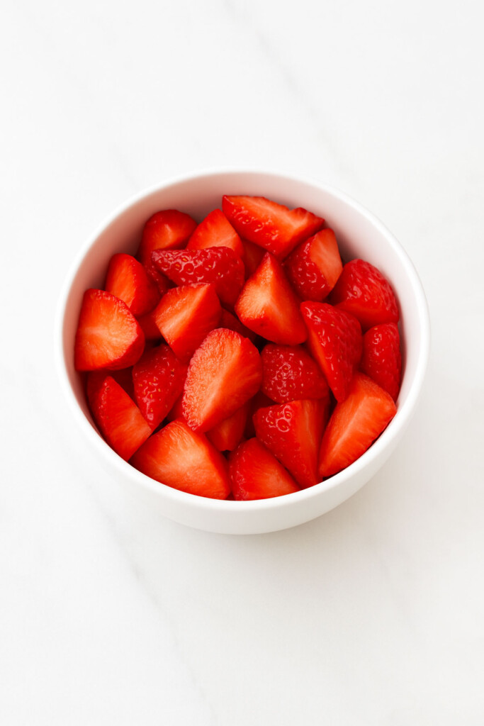 sliced strawberries in a white bowl on a marble counter.