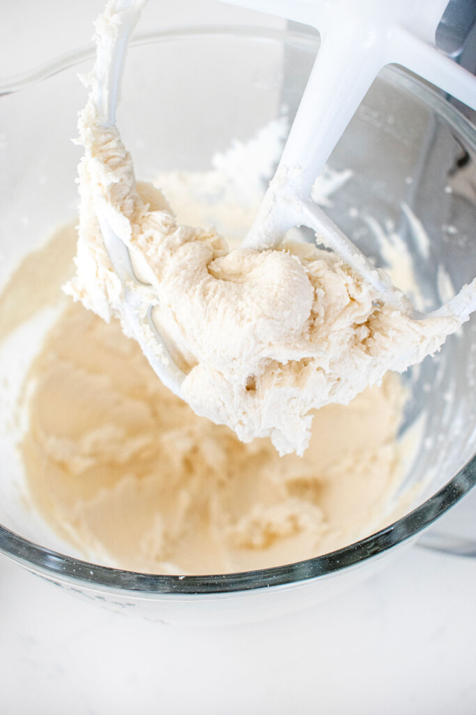 vanilla frosting in a glass bowl of a stand mixer on a white marble counter.