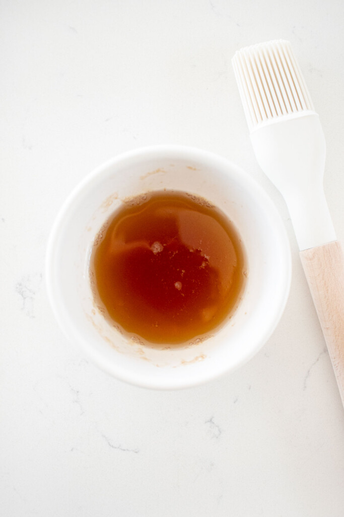 honey and water mixed together in a small bowl on a white marble counter with a pastry brush.