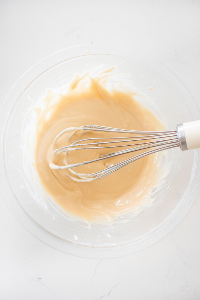 maple glaze being mixed with a whisk in a glass mixing bowl on a white marble counter.