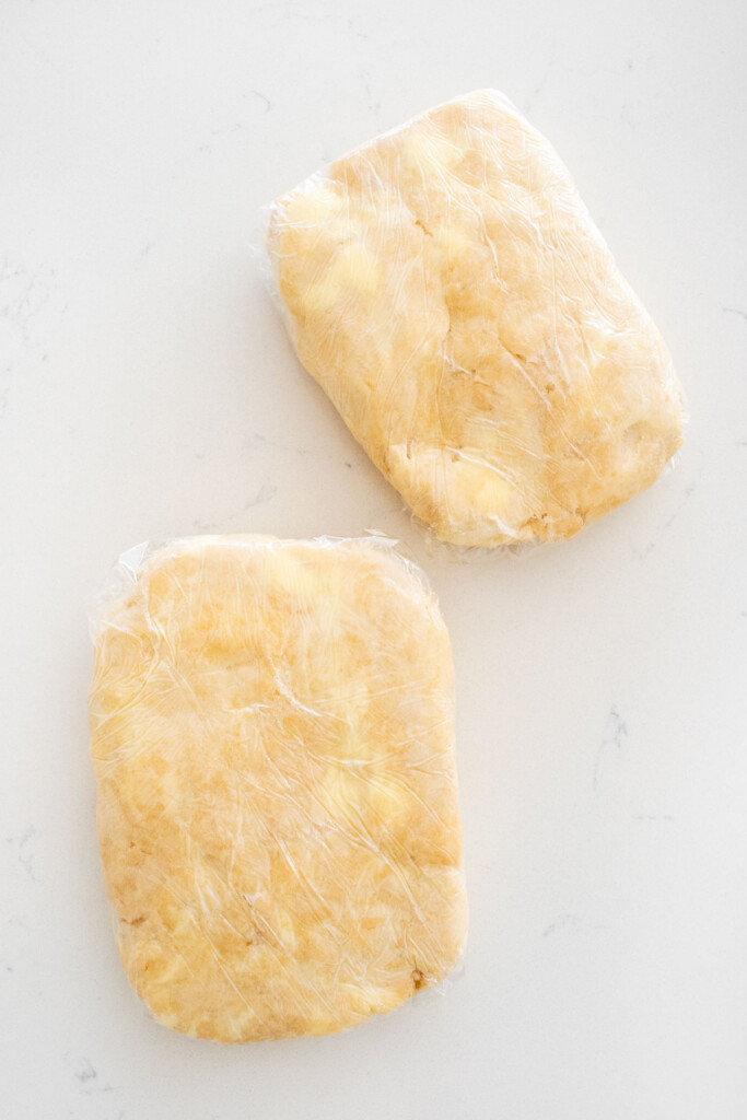 two rectangle pie dough pieces wrapped in plastic wrap on a white marble counter.
