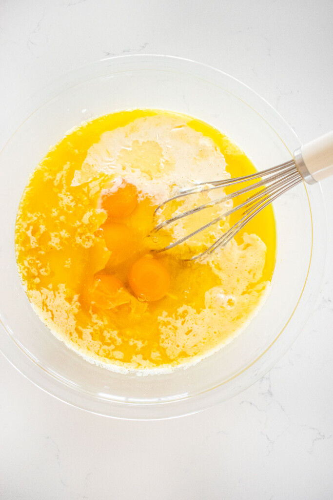 ingredients for eggnog waffles in a glass mixing bowl with a whisk on a white marble counter.