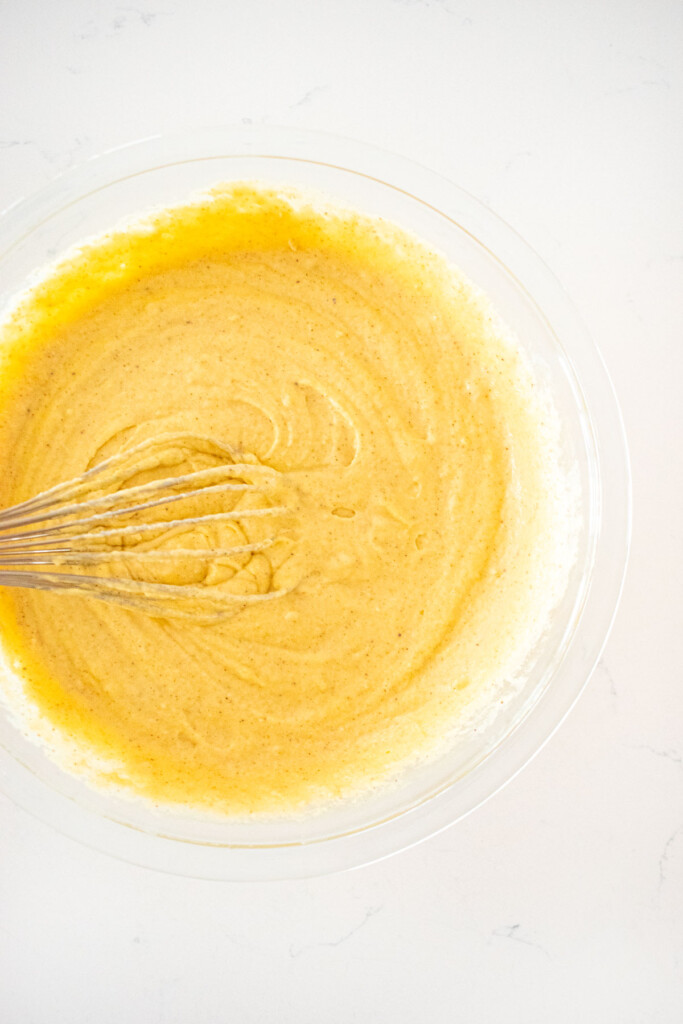 homemade eggnog waffle batter with a whisk in a glass mixing bowl on a white marble counter.