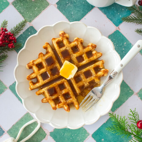 homemade eggnog waffles on a plate with butter and maple syrup on a green tile table.