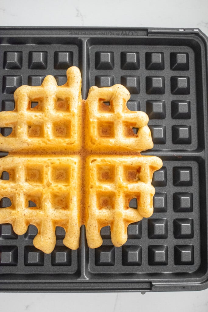 cooked eggnog waffle in a waffle iron on a white marble counter.