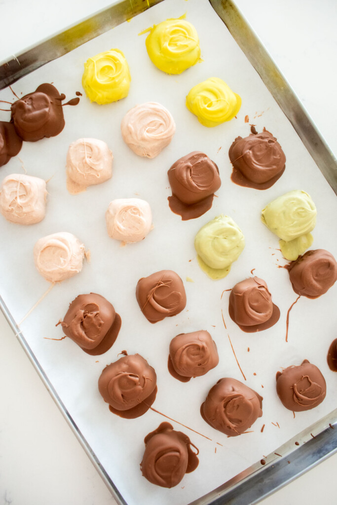 mint meltaways covered in milk chocolate and white chocolate that is light pink, green and yellow on a baking sheet on a marble counter.