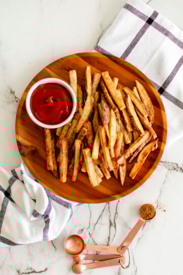 chinese five spice garlic french fries on a wood plate with a small dish of ketchup on a marble countertop.