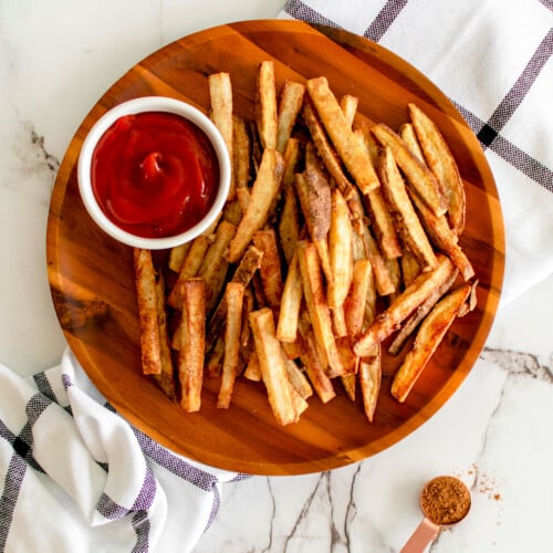 chinese five spice garlic french fries on a wood plate with a small dish of ketchup on a marble countertop.