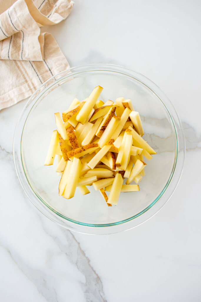 russet potatoes sliced into fries in a glass mixing bowl on a marble counter.