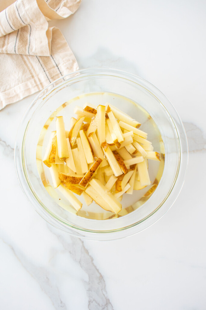 raw french fries soaking in water in a glass mixing bowl on a marble counter.
