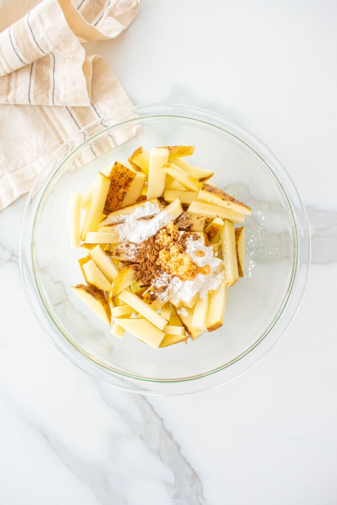 raw french fries with spices in a glass mixing bowl on a marble counter.