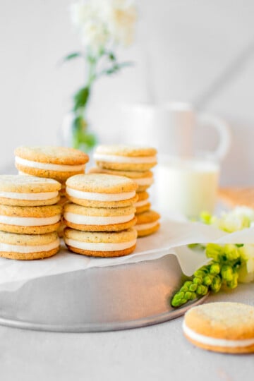 stacks of earl grey sandwich cookies on a silver platter on a grey counter with fresh flowers and a glass of milk.