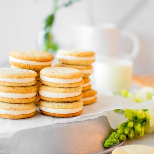 stacks of earl grey sandwich cookies on a silver platter on a grey counter with fresh flowers and a glass of milk.