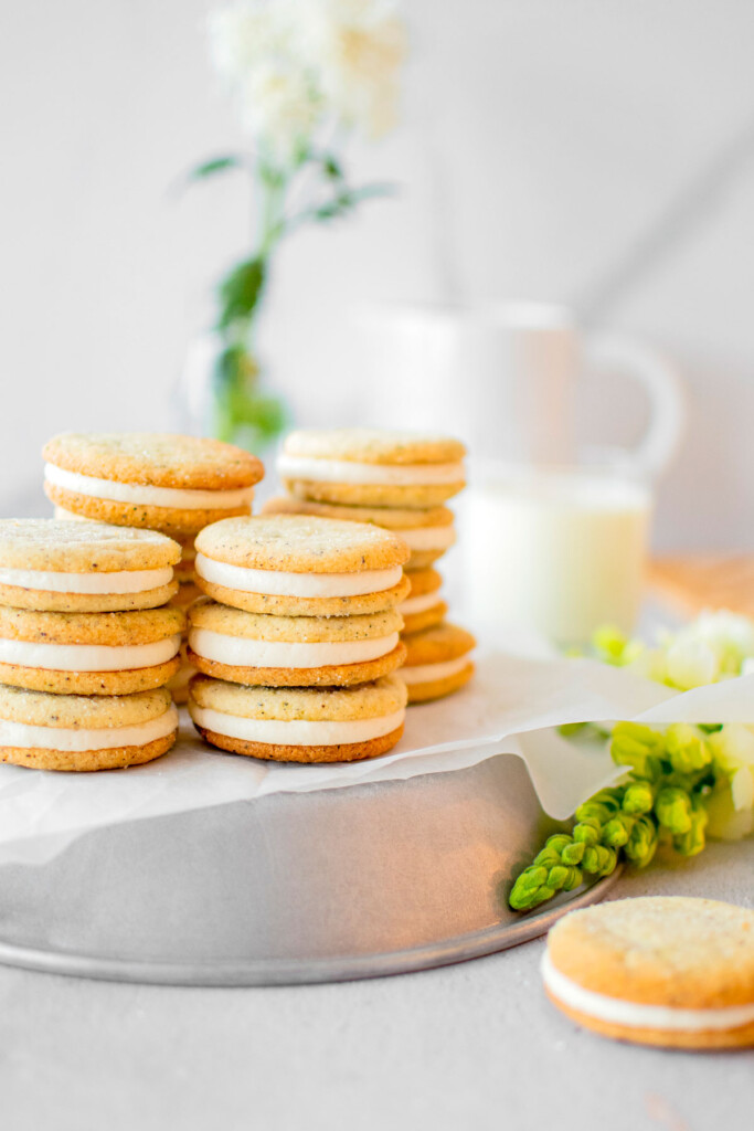 stacks of earl grey sandwich cookies on a silver platter on a grey counter with fresh flowers and a glass of milk.