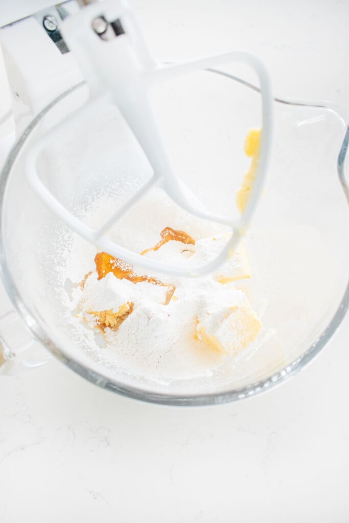 butter, powdered sugar, honey and vanilla in a glass stand mixing bowl on a white marble counter.