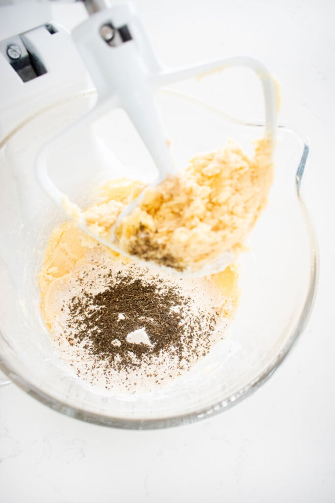 flour and earl grey tea added to creamed butter in a glass stand mixing bowl on a white marble counter.