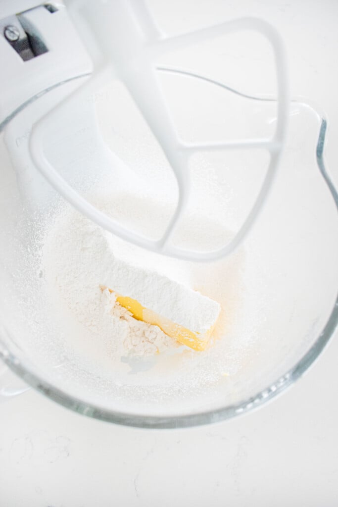 butter, powdered sugar and milk in a glass stand mixing bowl on a white marble counter.