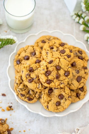 plate of chocolate chip cookies with malt powder on a table with a glass of milk and fresh white flowers.