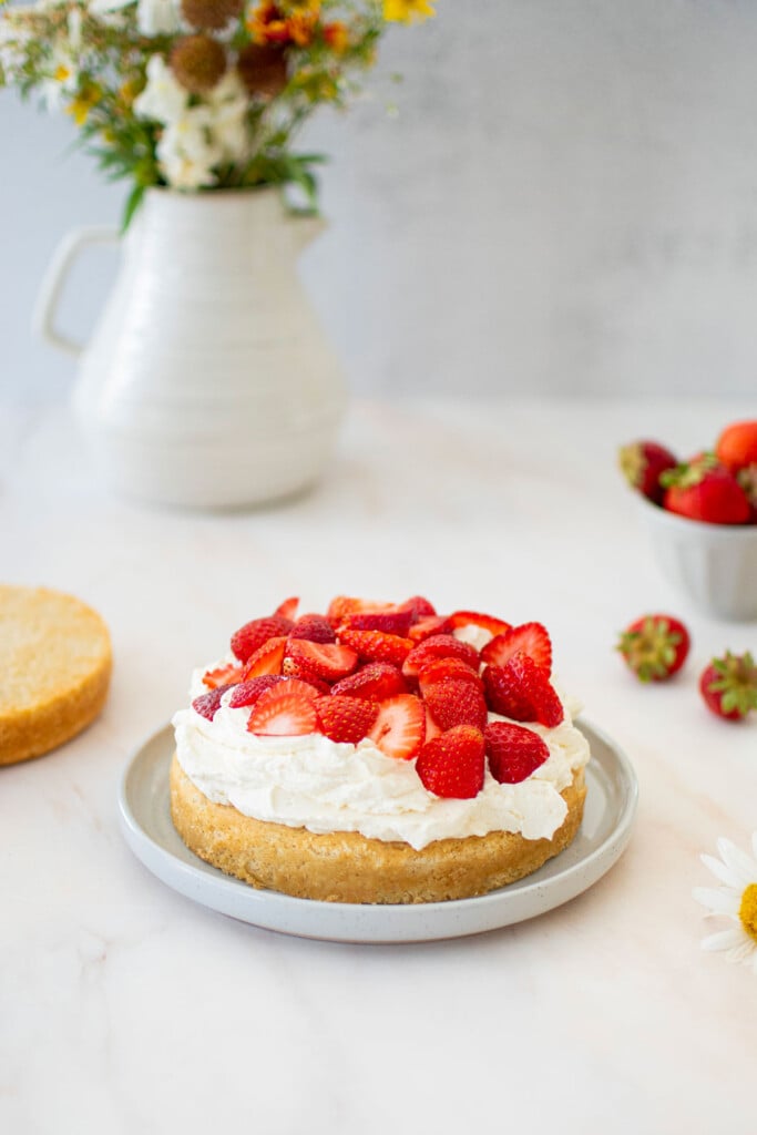 eggless strawberry shortcake on a cake plate with whipped cream and chopped strawberries on top on a marble counter.