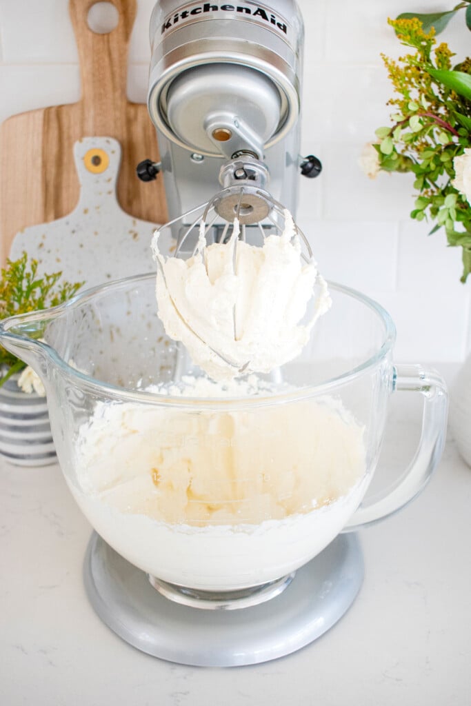 whipped cream in a glass mixing bowl of a stand mixer on a white marble countertop with flowers behind it.