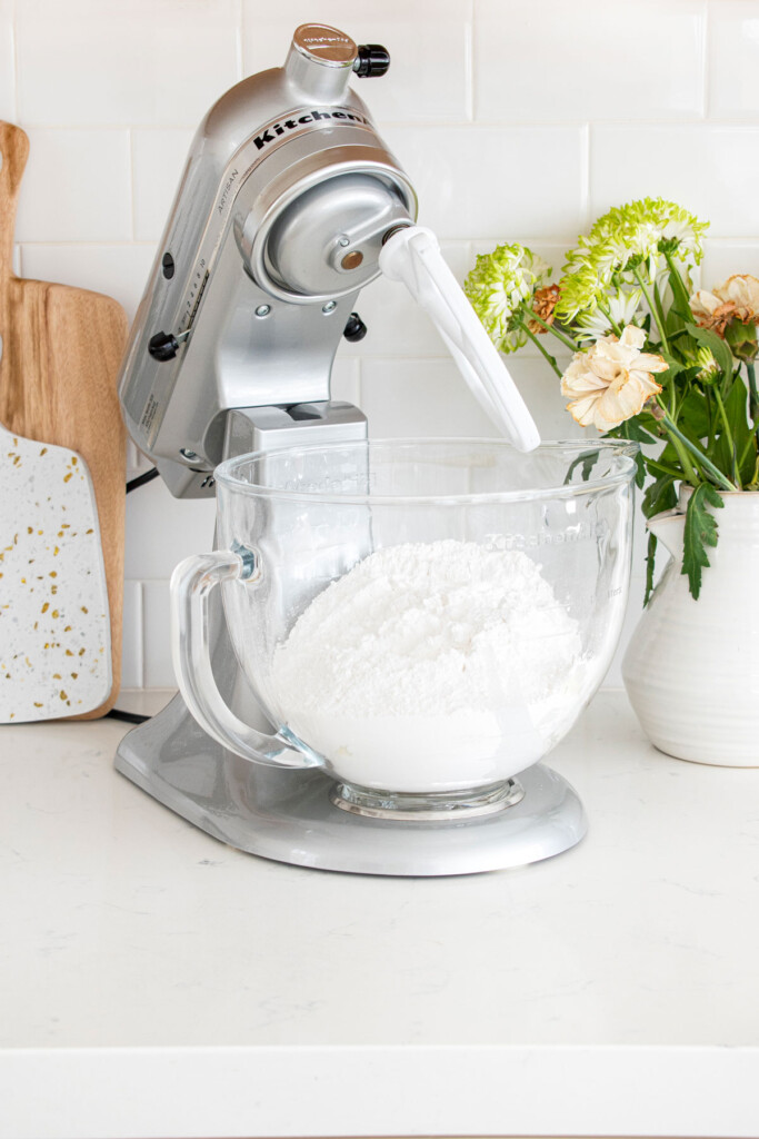 ingredients for vanilla frosting in a glass bowl of a stand mixer on a white marble counter with fresh flowers.