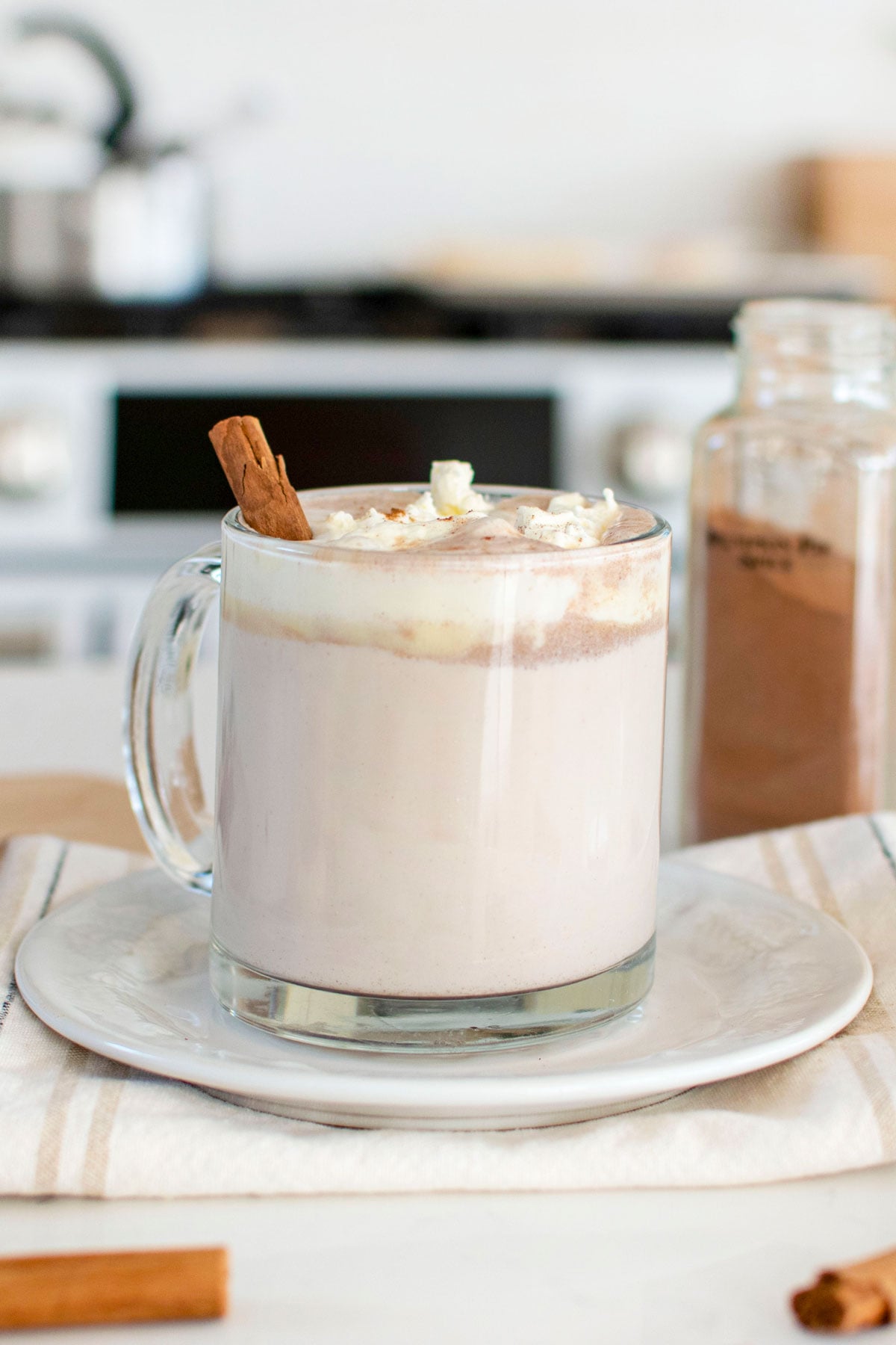 boozy pumpkin spice hot chocolate in a glass mug with whipped cream and a cinnamon stick on a plate on kitchen counter.