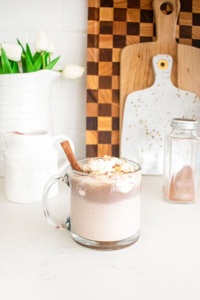 boozy pumpkin spice hot chocolate in a glass mug with whipped cream and a cinnamon stick on a white marble counter.