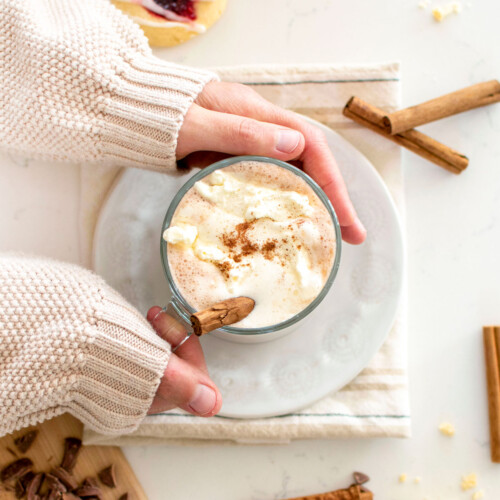 woman holding a glass mug with boozy pumpkin spice hot chocolate with whipped cream and a cinnamon stick.