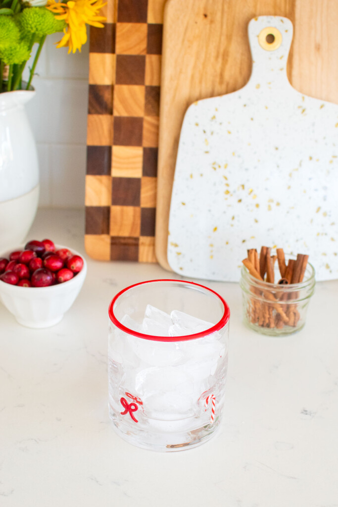 holiday punch with vodka being made in a holiday drink glass on a white marble counter with fresh cranberries and cinnamon sticks.