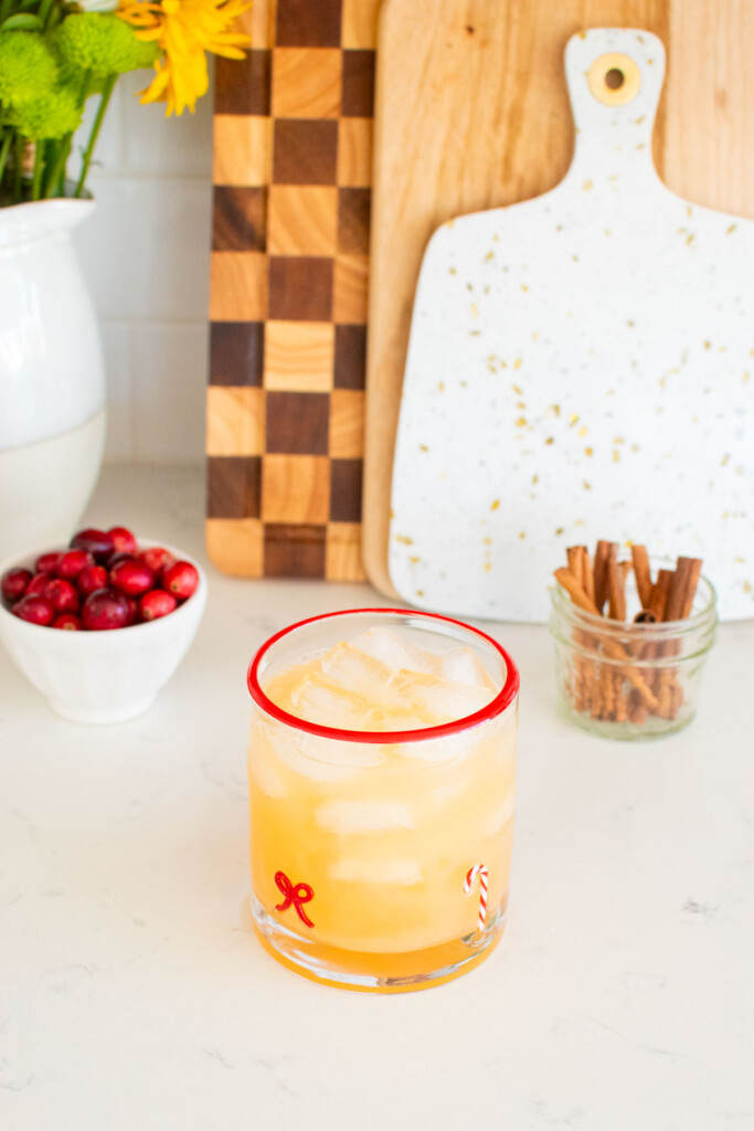 holiday punch with vodka being made in a holiday drink glass on a white marble counter with fresh cranberries and cinnamon sticks.