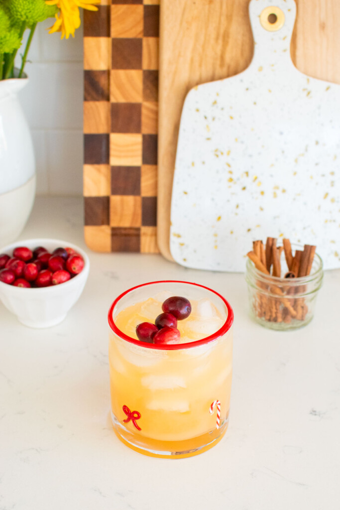 holiday punch with vodka being made in a holiday drink glass on a white marble counter with fresh cranberries and cinnamon sticks.