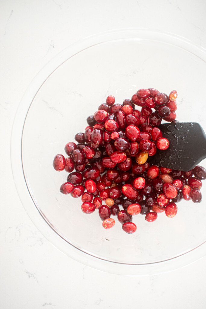 cranberries being coated in simple syrup in a glass mixing bowl on a white marble counter.